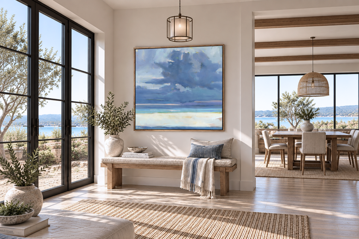 A large framed square art print depicting a stormy beach scene, displayed on a beige wall in a bright, modern living room with a wooden bench, plants, and a dining area in the background.